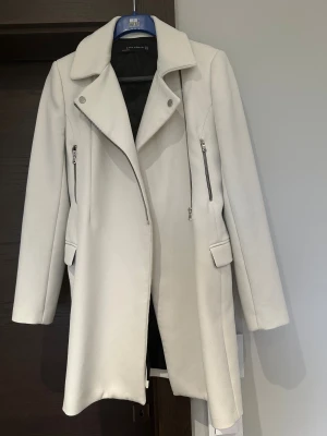 Vit kappa från Zara Woman med dragkedjor - Snygg vit kappa från Zara Woman i storlek M. Jackan har en stilren design med bred krage, silverfärgade dragkedjor och knappar framtill. Fickor med lock och raka, långa ärmar ger en clean look. Perfekt för dig som gillar minimalistisk stil.
