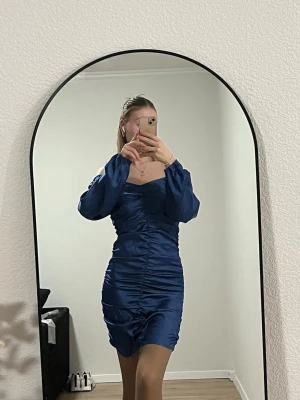 Marinblå omlottklänning med snörning - Säljer en marinblå kortklänning med snygg omlott-design och snörning längs hela framsidan. Klänningen har långa ärmar som kan bäras offshoulder och är gjord i ett glansigt, mjukt material som ger en lyxig känsla. Perfekt för dig som vill sticka ut med stil.