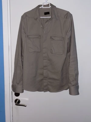 Beige overshirt från Rose & Born - Snygg beige overshirt från Rose & Born med två stora bröstfickor och klassisk krage. Tillverkad i mjuk bomull med lite elastan för extra komfort. Perfekt att slänga på över en t-shirt för en chill look.