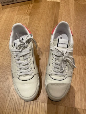 Emporio Armani vita sneakers med orange detalj size 38 - Snygga vita sneakers från Emporio Armani med orange detalj på hälen och svart sula. Skorna har snörning, rund tå och är tillverkade i skinn med präglad logga på sidan. Perfekt för dig som gillar stilrena och exklusiva sneakers med coola kontraster. Använda 3 gånger 