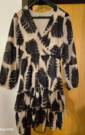 Beige och svart omlottklänning Look Up - Snygg kortklänning från Look Up med beige botten och svart abstrakt mönster. Klänningen har omlott-v-ringning, volangdetaljer och långa ärmar med resår vid ärmslut. Tillverkad i Italien, perfekt för dig som gillar trendiga och unika plagg.