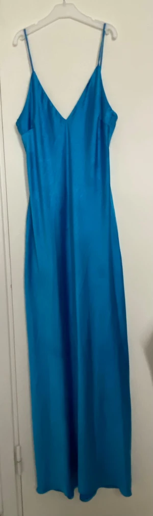 Blå långklänning med smala axelband - Snygg blå långklänning i satinliknande material med smala axelband och v-ringad hals. Klänningen har en elegant, enkel siluett som faller mjukt längs kroppen. Perfekt för dig som vill sticka ut med en färgstark look.