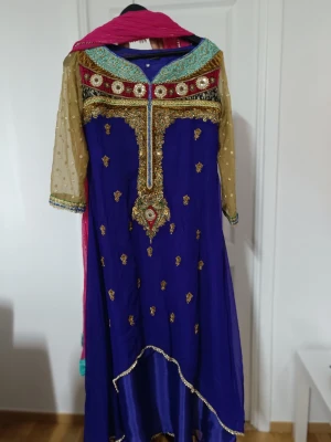 Blå långklänning med broderi och paljetter - Säljer en blå långklänning med guldiga broderier och paljettdetaljer, transparenta beige ärmar med prickar och en färgstark halsringning i turkos, rött och guld. Ingår även en rosa sjal med glittrande kant och en turkos-guldig mönstrad tygdel. Perfekt för dig som gillar färg och detaljer!