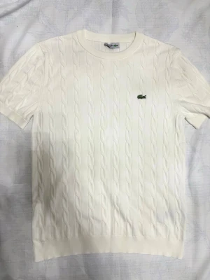 Lacoste Tröja - Vit kabelstickad tröja från Lacoste med korta ärmar och rund hals. Toppen har ett diskret kabelstickat mönster och den klassiska gröna krokodilloggan broderad på bröstet. Perfekt för dig som gillar stilren och oldschool design.