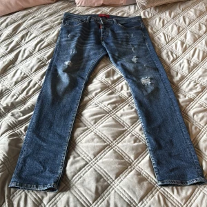 Blå Dsquared2 jeans med slitningar - Snygga blå jeans från Dsquared2 med klassisk femficksmodell och coola slitningar på låren och knäna. Jeansen har rak passform och är tillverkade i jeansmaterial med tydliga detaljer som röd logotagg och läderpatch bak.
