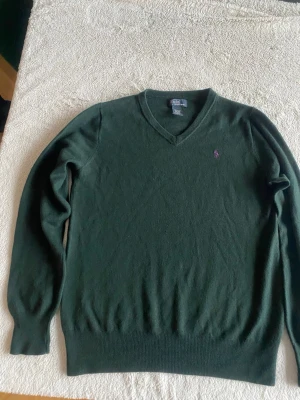 Mörkgrön v-ringad tröja Polo Ralph Lauren - Mörkgrön v-ringad tröja från Polo Ralph Lauren i 100% merinoull. Klassisk design med lila broderad logga på bröstet och ribbade muddar vid ärmslut och nederkant. Perfekt för dig som gillar stilrena och tidlösa plagg.