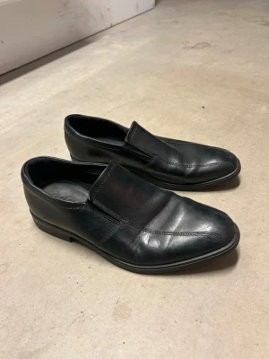Svarta loafers från Ecco i skinn - Stilrena svarta loafers från Ecco i äkta skinn. Skorna har en klassisk design med rund tå och platt sula, perfekt för dig som gillar en clean och tidlös look. Passar till både jeans och kostymbyxor. Snygga sömmar och diskret branding på insidan. Finns några repor här och där. 