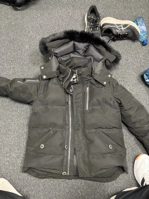 Svart Moose Knuckles dunjacka S - Svart dunjacka från Moose Knuckles med fet broderad hockeypatch på insidan. Jackan har pufferstil, dragkedja, knappstängning och fickor med lock. Huva med svart fuskpäls och coola detaljer som metallöglor och innerfickor. Perfekt för kalla dagar.