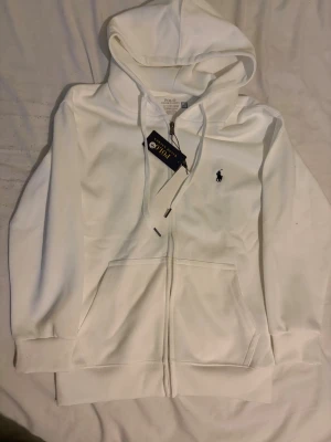 Vit hoodie från Polo Ralph Lauren - Vit hoodie från Polo Ralph Lauren med klassisk svart broderad logga på bröstet. Slim fit-modell med dragkedja, huva med snörning och två fickor framtill. Tillverkad i mjuk bomull som känns skön mot huden. Perfekt för en clean och stilren look.