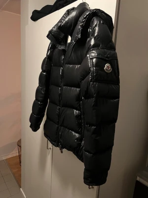 Svart Moncler Maya strl4 - Säljer en svart pufferjacka från Moncler med glansig finish och huva. Jackan har Moncler-logga på ärmen, dragkedja framtill och en cool serietidningsetikett på insidan. Perfekt för kalla dagar och har en riktigt schysst streetstil.