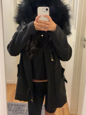 Svart parkajacka med fuskpäls från DM - Säljer en svart parkajacka med stor huva kantad av fluffig svart fuskpäls. Jackan har dragkedja, knappar och snygga detaljer i guld. Fickor på sidorna och justerbara band vid midjan. Perfekt för kyliga dagar och riktigt snygg till streetwear.