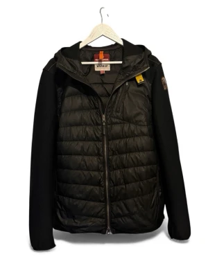 Parajumpers jacka (XL) - Svart pufferjacka från Parajumpers i storlek XL. Jackan har huva, quiltad framsida och rygg, samt släta ärmar med patch på axeln. Materialet är polyester och syntet, med dragkedja framtill och en extra ficka med zip på ryggen. Perfekt för kyliga dagar. Jackan har ett jättelite hål på vänster ficka, och sömmarna har lossnat jättelite på baksidan av jackan. För fler bilder på jackan eller funderingar är det bara att höra av dig.