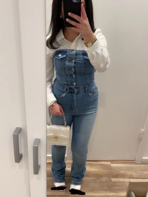 Jeans jumpsuit / byxdress – snygg och stilren - Säljer en snygg ljusblå jeansbyxdress med hög midja och raka ben. Byxdressen har flera silverfärgade knappar framtill och klassiska jeanssömmar. Perfekt för dig som gillar en cool och trendig denimlook.