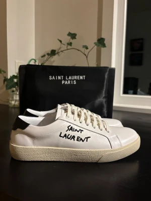 Saint Laurent vita sneakers med svart detalj - Snygga vita sneakers från Saint Laurent med handskriven logga på sidan och svart detalj vid hälen. Skorna har rund tå, platt sula och klassisk snörning. Tillverkade i skinn med en stilren och minimalistisk design som ger en lyxig vibe. Skorna är ej använda endast testade