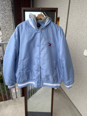 Ljusblå bomberjacka Tommy Jeans - Snygg ljusblå bomberjacka från Tommy Jeans med broderad logga på bröstet och ryggen. Jackan har ribbade muddar med vita ränder, tryckknappar framtill och en grå huva i sweatshirtmaterial. Perfekt för dig som vill ha en chill och sportig stil.