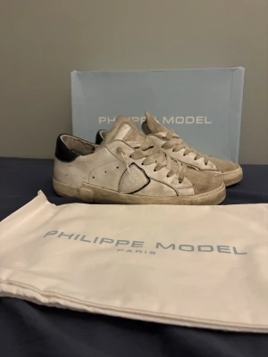 Philippe Model vita sneakers i skinn - Snygga vita sneakers från Philippe Model med beige mockadetaljer och svart häl. Klassisk låg modell med snörning och rund tå. Tillverkade i skinn och mocka, med logga på sidan och plös. Kommer med originalkartong och dustbag.