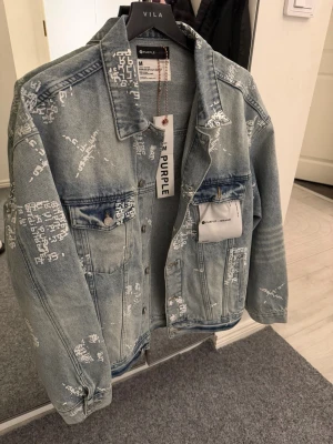 Ljusblå jeansjacka från Purple Brand - Säljer en ljusblå jeansjacka från Purple Brand med coola vita tryck över hela jackan. Klassisk modell med krage, knappar framtill och två bröstfickor. Jackan har en loose passform och är tillverkad i slitstarkt bomullsmaterial. Perfekt för dig som gillar streetstyle.