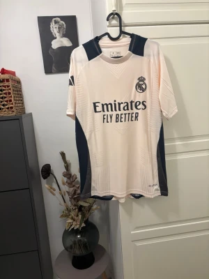 Adidas Real Madrid trainer jersey player version  - Snygg Real Madrid matchtröja från Adidas i slim fit, storlek M. Tröjan är ljusbeige med marinblå detaljer och har HEAT.RDY-teknologi för extra komfort. Ikoniskt klubbmärke och Adidas-logga på ärmen. Perfekt för dig som älskar fotboll och vill ha en stilren look.