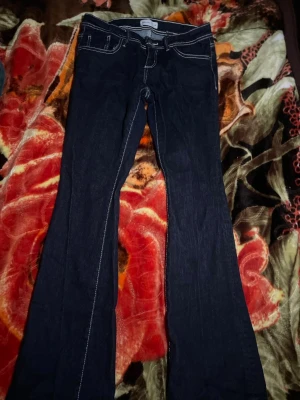 Mörkblå bootcut jeans med kontrastsömmar - Snygga mörkblå jeans med bootcut passform och vita kontrastsömmar. Klassisk femficksmodell med bälteshällor och knappstängning. Perfekta för dig som gillar en retro vibe och vill ha ett par jeans som sticker ut lite extra.