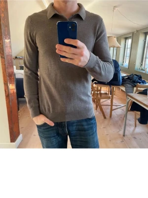 Brun pikétröja i ull från John Henric - Snygg brun långärmad pikétröja från John Henric i mjuk ull. Tröjan har klassisk krage, två knappar framtill och ribbade muddar vid ärmslut och nederkant. Perfekt för dig som gillar stilrena och enkla plagg med en chill vibe.