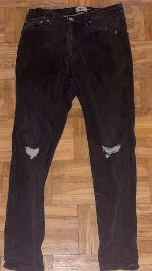 Svarta jeans med slitna knän - Svarta jeans från 157 med snygga slitningar på båda knäna. Modellen är rak och har klassiska fem fickor samt knapp och dragkedja framtill. Jeansen har en cool, mörk tvätt och passar perfekt för en avslappnad streetstil. helt oanvända
