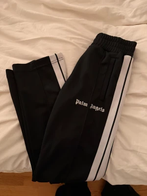 Palm Angels byxor - Svarta byxor från Palm Angels med vita sidoremsor och logotyp broderad på låret. Byxorna har elastisk midja, raka ben och är tillverkade i ett mjukt syntetmaterial som ger en skön och avslappnad vibe. Perfekta för chill eller streetwear.