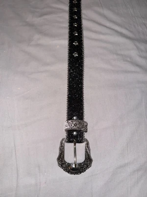 Svart glittrig bälte med stjärnor - Snyggt svart bälte med glittrig yta och silverfärgade stjärnformade nitar längs hela bandet. Spännet och ändarna har detaljerade, blommiga mönster i silver. Perfekt för att ge outfiten en extra edge och western-vibe. Helt oanvänt då jag köpte det till en fest men paketet kom inte i tid. 