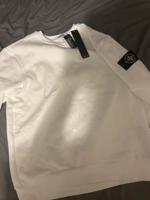 Vit sweatshirt från Stone Island - Vit sweatshirt från Stone Island med klassisk rund hals och ribbade muddar. Tröjan har den ikoniska Stone Island-patchen på vänster ärm och är tillverkad i mjuk bomull. Perfekt för dig som gillar clean och stilren streetwear. Perfekt som jul present eller för vintern 