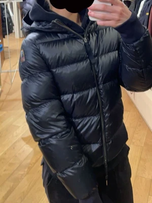 Svart dunjacka Parajumpers - Säljer en svart dunjacka från Parajumpers med huva och quiltad design. Jackan har dragkedja framtill och ribbade muddar vid ärmslut. Insidan är ljus och jackan har en patch med logga på ärmen. Har storlek L då den kändes liten i storleken och för att jag skulle kunna ha tjockare tröjor under. Sitter mer som en M. Nypris runt 6000-6300. Köpt från johnells. Hittar inte den på sidan.