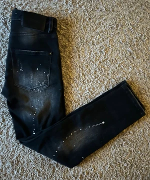 Svarta Dsquared2 Jeans - Svarta jeans från Dsquared2 med coola slitningar och vita färgstänk. Jeansen har klassisk femficksdesign, knappgylf och slim passform. Materialet är jeans med stretch för extra komfort. Perfekta för dig som gillar en edgy och trendig look.