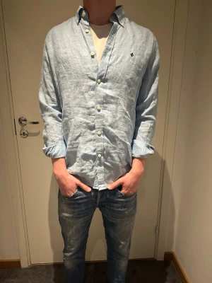 Ljusblå linneskjorta Morris - Säljer en ljusblå skjorta från Morris i svalt linne. Skjortan har klassisk krage, knappar framtill och en broderad logga på bröstet. Långärmad modell med avslappnad passform, perfekt till jeans eller shorts.pris kan diskuteras vid snabb affär