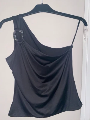 Svart oneshoulder topp med metallring - Svart oneshoulder topp i mjukt, glansigt material med en cool silverfärgad metallring vid axeln. Toppen har draperad front och en smal axelrem på ena sidan. Perfekt för dig som gillar edgy och stilrena plagg med unik detalj.