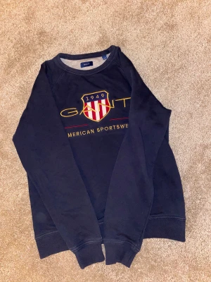 Mörkblå sweatshirt från GANT - Mörkblå sweatshirt från GANT med broderad logga och sköld framtill i gult, rött och vitt. Tröjan har rund halsringning, långa ärmar och ribbade muddar. Tillverkad i mjuk bomull, perfekt för en chill och sportig stil.