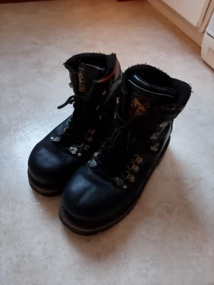 Svarta kängor Ten Points strl 44 - Stabila svarta kängor från Ten Points med grov sula och snörning. Skorna har detaljer i brunt på hälen och metallhakar för snörningen. Ovandelen är i skinn och insidan är fodrad för extra komfort. Perfekt för dig som gillar robust stil och funktion.