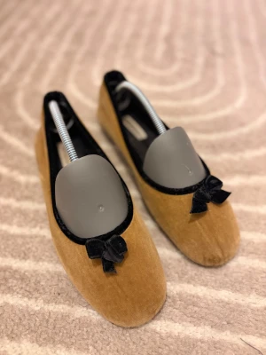 Bruna ballerinaskor i sammet från Hush Puppies - Snygga ballerinaskor i brun sammet från Hush Puppies med svarta sammetsdetaljer och en söt rosett framtill. Skorna har rund tå och platt sula, perfekt för dig som gillar en klassisk och chic look. Mjukt material och stilren design. Passar som innetofflor 