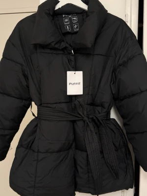 Svart pufferjacka - Svart pufferjacka från Puffit i storlek XS. Jackan har bälte i midjan och är helt ny. Säljer eftersom jag råkade köpa fel storlek. Köpt för 650.
