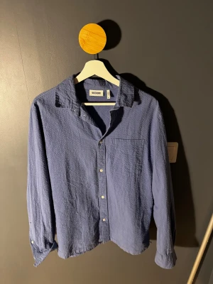 Blå skjorta från Weekday i bomull - Säljer en blå skjorta från Weekday i storlek M. Skjortan är tillverkad i ett strukturerat bomullstyg, har klassisk krage, bröstficka och knäppning framtill. Perfekt för en chill och stilren look.