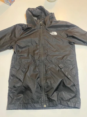 Svart vindjacka från The North Face - Svart vindjacka från The North Face med huva och hel dragkedja framtill. Jackan har två snedställda fickor och logga på bröstet. Materialet är lätt och syntetiskt, perfekt för blåsiga dagar. Klassisk passform och långärmad design.