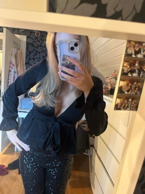 Svart omlottblus från Bik Bok - Säljer en svart omlottblus från Bik Bok med djup v-ringning och knyt i midjan. Blusen har långa ärmar med lätt puff och resår vid ärmslut. Materialet är glansigt och känns lyxigt, perfekt för att lyfta din outfit. Supersnygg att styla med glittriga byxor eller jeans.