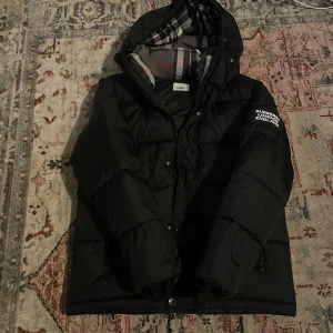 Svart Burberry pufferjacka med huva/också en vestjacka - Svart pufferjacka från Burberry med avtagbara ärmar, huva och klassiskt rutigt foder. Jackan har en vit Burberry London England-logga på ärmen och dragkedja framtill. Perfekt för kalla dagar och riktigt snygg streetstil.