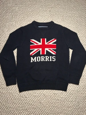 Mörkblå stickad Morris tröja Union Jack - Snygg mörkblå stickad tröja från Morris i storlek medium. Framsidan har ett stort Union Jack-tryck i rött, vitt och blått samt Morris-logga under. Tröjan har rund hals, ribbade muddar och är långärmad. Perfekt för dig som gillar preppy stil och tydliga detaljer. Nytt skick utan prislapp och påse.