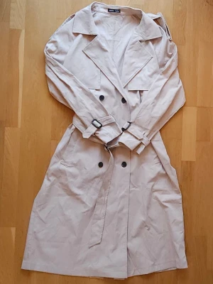 Beige trenchcoat - Snygg beige trenchcoat med klassisk krage, dubbelknäppning och bälte i midjan. Jackan har långa ärmar med spännen vid handlederna och svarta knappar framtill. Perfekt för en stilren och tidlös look. Är något liten i storleken och skulle säga att trenchcoaten är mer en L än XL. Har aldrig blivit använd.