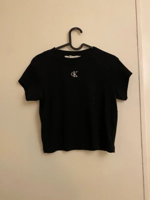 Svart t-shirt från Calvin Klein - Svart t-shirt från Calvin Klein med klassisk rund hals och korta ärmar. Framtill finns en liten vit CK-logga broderad på bröstet. Modellen är croppad och har en stilren, enkel design som funkar till många olika looks.