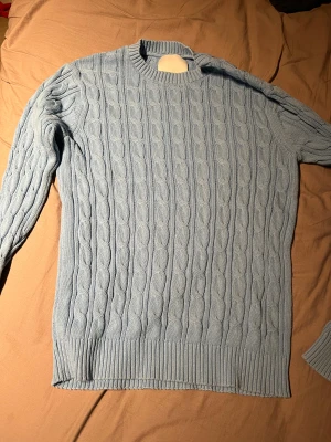 Ljusblå kabelstickad tröja - Snygg ljusblå kabelstickad tröja med rund hals och långa ärmar. Tröjan har ett klassiskt flätmönster och ribbade muddar vid ärmslut och nederkant. Perfekt för dig som gillar stilrena och mysiga plagg med lite preppy vibe.