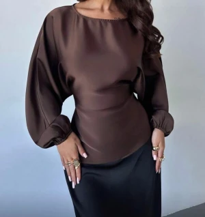 Brun omlott blus med ballongärmar - Säljer en brun blus i satinliknande material med ballongärmar och rund hals. Toppen har en snygg omlott-effekt med knyt i ryggen som framhäver midjan. Perfekt för dig som gillar stilrena och trendiga plagg med lite extra volym i ärmarna.