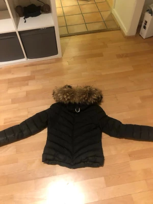 Svart pufferjacka med päls från Holies - Svart pufferjacka från Holies med snyggt leopardmönstrat foder och avtagbar päls på huvan. Jackan har guldfärgade dragkedjor, quiltad design och långärmade armar. Perfekt för dig som vill ha en varm och cool look under vintern.