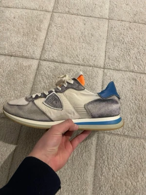 Philippe Model sneakers med blå detalj - Säljer ett par Philippe Model sneakers i vitt och grått med inslag av blått och orange. Skorna har snörning, rund tå och platt sula. Materialmix av mocka och textil, med märkets logga på sidan. Perfekt för dig som gillar stilrena och sportiga  sneakers. Funkar perfekt för hela året! Kom med prisförslag🤩👍