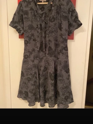 Grå blommig kortklänning med knyt - Säljer en grå kortklänning med blommigt mönster och knytband vid halsen. Klänningen har korta ärmar och ett mjukt, svängigt fall. Perfekt för dig som gillar detaljer och mönster. Passar dig som vill ha en unik och cool look.