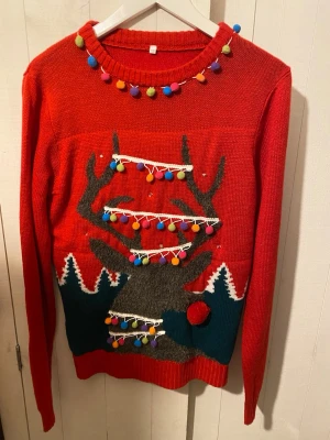 Röd jultröja med lampor - Riktigt färgstark jultröja i rött med ett stort renmotiv framtill, dekorerad med färgglada bollar och inbyggda lampor som lyser. Tröjan har ribbade muddar, lång ärm och detaljer i grönt, vitt och grått. Perfekt för dig som vill sticka ut på julmyset! Passar från XS-M