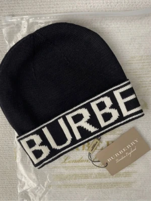 Svart Burberry mössa med vit logga - Snygg svart mössa från Burberry med bred vit kant och stor stickad logga framtill. Mössan är stickad och har en klassisk form som passar perfekt till streetwear och vinteroutfits. Materialet är mjukt och håller dig varm under kalla dagar.